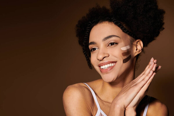A young African American woman with a bright smile stands against a beige backdrop, showcasing makeup swatches on her cheek