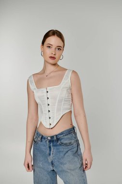 Güzel genç bir kadın, açık gri bir arkaplana karşı beyaz bir korsenin içinde güven içinde poz veriyor..