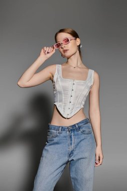 Beyaz korseli ve kot pantolonlu genç bir kadın gri bir arkaplana karşı güvenle poz veriyor..