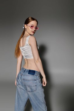 Uzun kızıl saçlı genç bir kadın gri arka planda poz veriyor. Üzerinde kot pantolon ve beyaz bluz var..