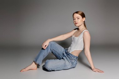 A young woman poses in a white corset and jeans against a gray backdrop. clipart