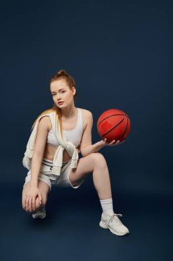 Young woman in white sports bra, shorts, holding basketball against vivid blue. clipart