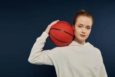 Omzuna basketbol topu dayamış genç bir kadın mavi bir zemine karşı poz veriyor..