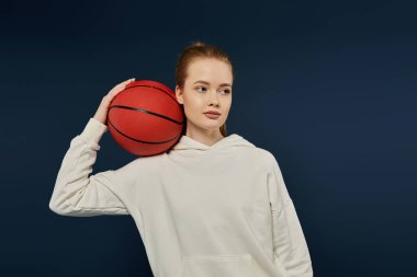 Genç bir kadın mavi arka planda basketbol topuyla poz veriyor..