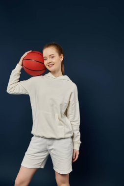 Genç bir kadın basketbol topuyla mavi arka planda poz veriyor..