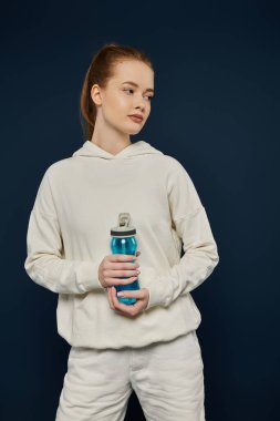 Beyaz kapüşonlu ve pantolonlu genç bir kadın mavi zemine karşı su şişesiyle poz veriyor..