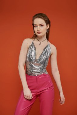 Woman poses in silver top and hot pink pants against orange backdrop. clipart