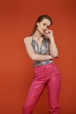 Gümüş rengi bluzlu ve pembe deri pantolonlu genç bir kadın canlı turuncu bir fonda poz veriyor..