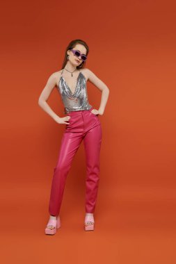 Young woman in pink pants and silver top poses with hands on hips against bright orange backdrop. clipart