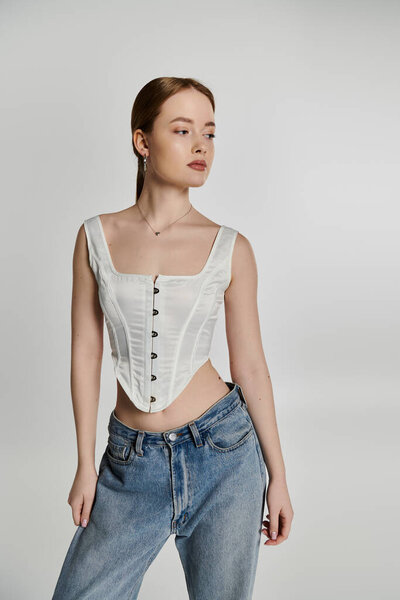 A young woman stands against a white backdrop, wearing a white corset and blue jeans.