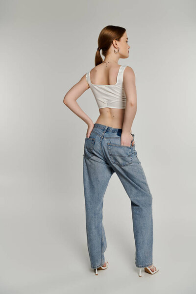 A young woman with her back to the camera poses in light-wash denim jeans and a white crop top.