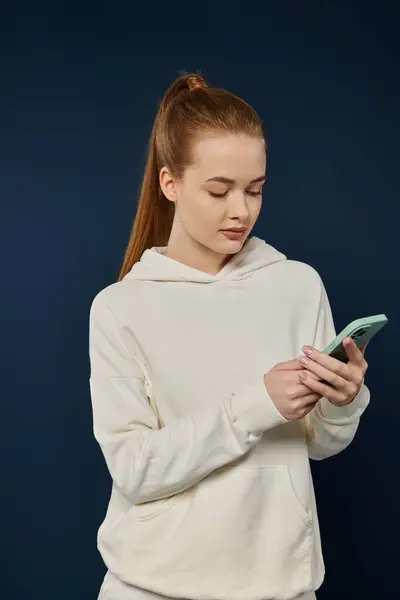 stock image A young woman in a white hoodie looks intently at her phone against a vibrant blue backdrop.