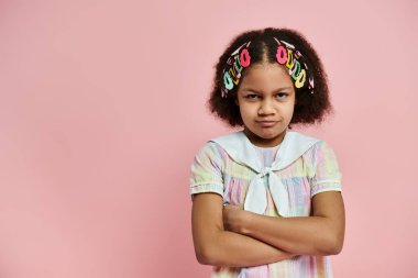 A young African American girl with hair clips stands with her arms crossed on a pink background. clipart