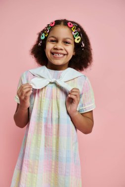 Renkli saç tokalarıyla genç bir Afrikalı Amerikalı kız pembe arka planda duran renkli bir elbise içinde ışıl ışıl gülümsüyor..