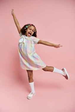 Renkli elbiseli ve tokalı genç bir Afrikalı Amerikalı kız pembe bir arka planda duruyor, neşeli bir poz veriyor..