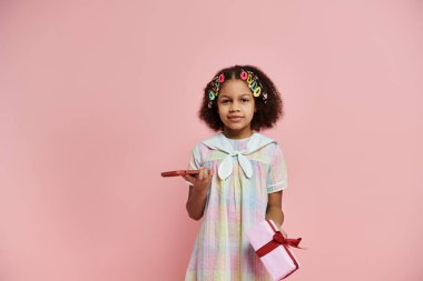 A young African American girl in a colorful dress holds a pink gift box and a red phone on a pink background. clipart