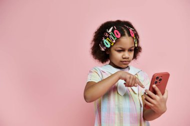 Renkli saç tokalarıyla genç bir Afrikalı Amerikalı kız pembe bir arka planda duruyor ve akıllı telefonuna bakıyor..