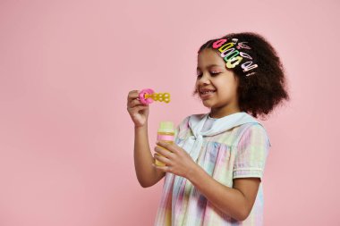 A young African American girl in a colorful dress blows bubbles with a pink wand against a pink background. clipart