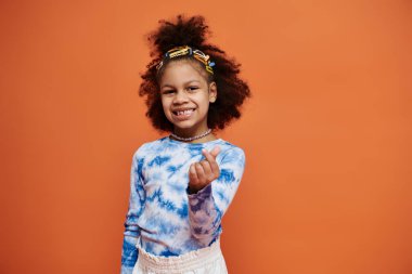 A young African American girl with curly hair and hair clips smiles at the camera while wearing a blue tie-dye shirt. clipart