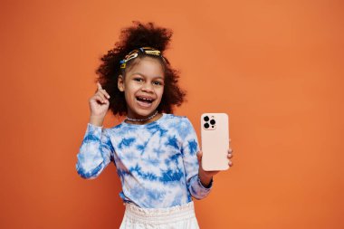 Renkli saç tokaları ve modaya uygun kıyafetleri olan genç bir Afrikalı Amerikalı kız akıllı telefonuyla turuncu arka planda poz veriyor..