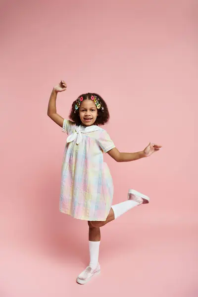 stock image A joyful African American girl in a colorful dress and hair clips smiles for the camera against a pink background.