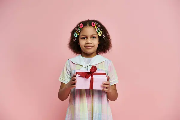 stock image A young African American girl in a colorful dress stands on a pink background, holding a gift box with a red ribbon.