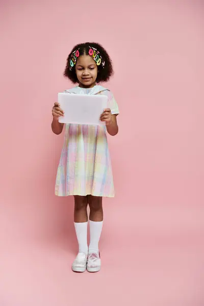 stock image A young African American girl in a colorful dress stands on a pink background and looks intently at a tablet.