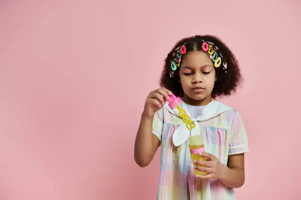 stock image A cute African American girl in a pink and white dress and colorful hair clips holds a bottle in her hand.