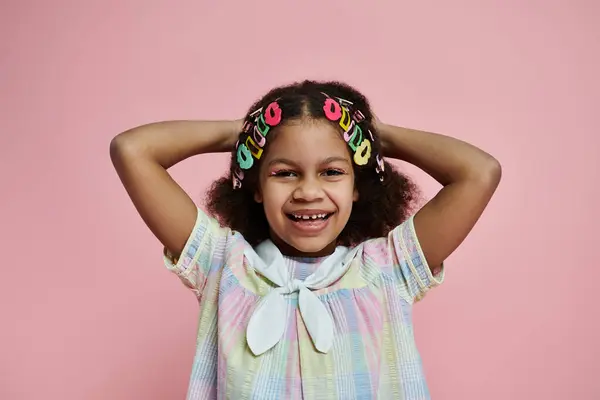 stock image A young African American girl with hair clips stands in a colorful dress against a pink backdrop, radiating joy and happiness.