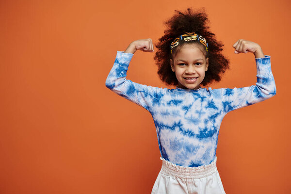 Молодая афроамериканка со стильными заколками для волос и рубашкой-галстуком демонстрирует свои мышцы на фоне оранжевого цвета.