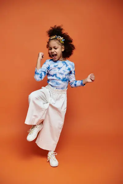 stock image A young girl with stylish hair clips stands confidently in a trendy outfit, striking a joyful pose against an orange backdrop.