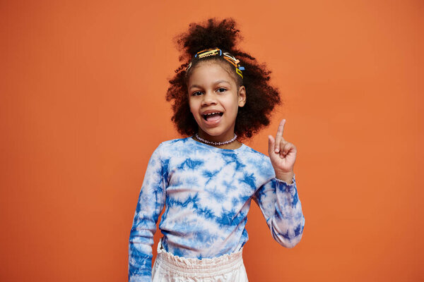 Молодая афроамериканка с красочными заколками для волос стоит в модном наряде на оранжевом фоне, указывая пальцем вверх..