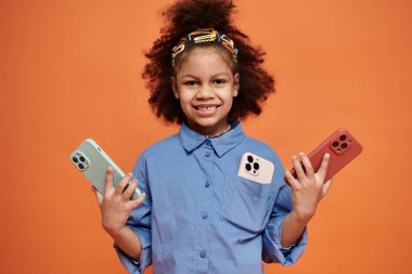 Mavi gömlekli ve saç tokalı Afro-Amerikalı bir kız elinde iki akıllı telefonla turuncu bir fonda duruyor ve gülümsüyor..