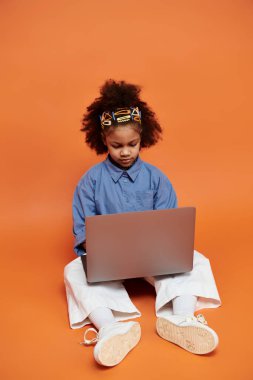 Renkli saç tokaları olan şık bir Afrikalı Amerikalı kız turuncu bir arka planda oturuyor ve dizüstü bilgisayar kullanıyor..