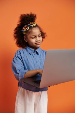 Renkli saç tokaları olan şık bir Afro-Amerikalı kız, modaya uygun bir kıyafet içinde dizüstü bilgisayarını turuncu arka planda kullanıyor..