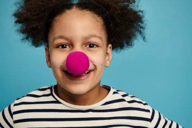 Kıvırcık saçlı, palyaço burnu takan ve mavi arka planda gülümseyen Afro-Amerikan bir kızın yakın plan portresi..