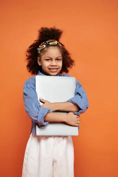 stock image A young African American girl in a trendy outfit smiles brightly while holding a laptop against an orange background.