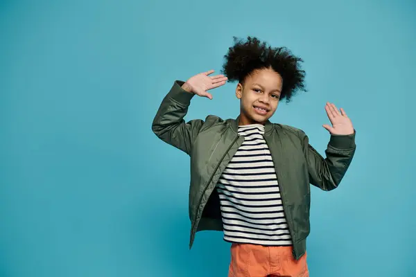 stock image Confident young African American girl in green bomber jacket and striped shirt stands against bright blue background with curly hair.