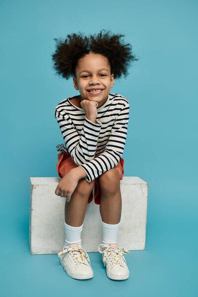Young African American girl sits on white platform with curly hair, striped shirt, red shorts, and white sneakers against blue background.