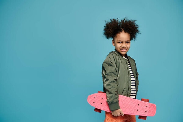 Молодая афроамериканка с вьющимися волосами улыбается, держа розовый скейтборд на синем фоне.