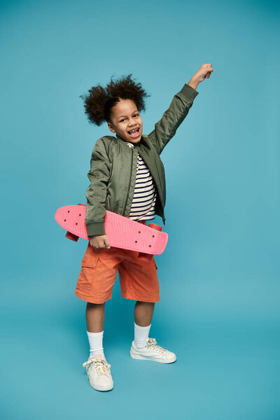 Молодая афроамериканка с вьющимися волосами ярко улыбается, держа розовый скейтборд на фоне синего фона..
