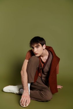 Süslü bir adam stüdyoda poz veriyor, kahverengi bir süveter ve pantolon giyiyor..