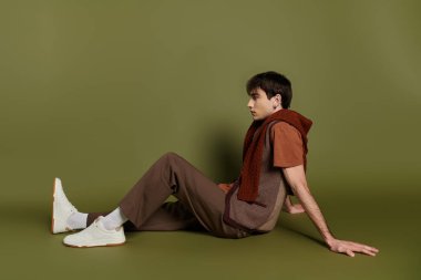 Kahverengi pantolonlu, beyaz ve kahverengi yelekli, kestane rengi eşarp yeşil arka planda poz veriyor.