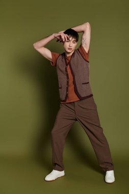 Tarz sahibi bir adam yeşil bir zemine karşı yelek ve kahverengi pantolonla poz verir..