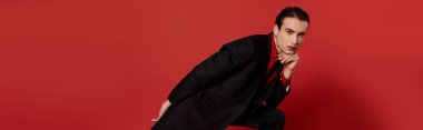 Siyah elbiseli bir adam kırmızı arka planda poz veriyor..