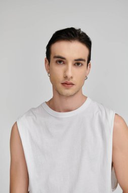 Beyaz kolsuz gömlekli genç bir adam minimalist bir arkaplana karşı poz veriyor..