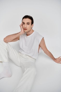 Beyaz kolsuz gömlekli ve beyaz pantolonlu genç bir adam beyaz bir fona karşı poz veriyor..