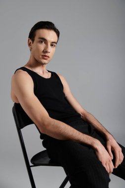 A man wearing a black tank top poses on a black chair against a grey backdrop. clipart