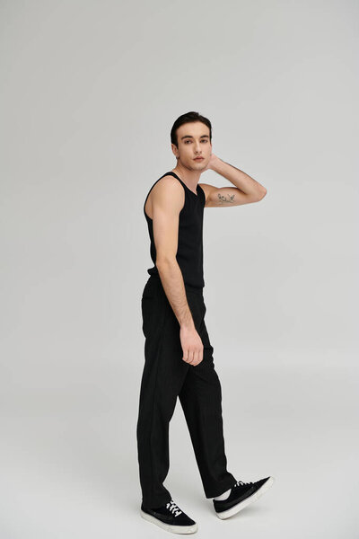 A man in black tank top and trousers poses against a gray backdrop.