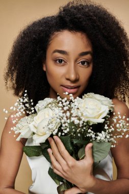 A beautiful African American woman with natural makeup holds a bouquet of white roses and baby breath flowers in front of a beige background.